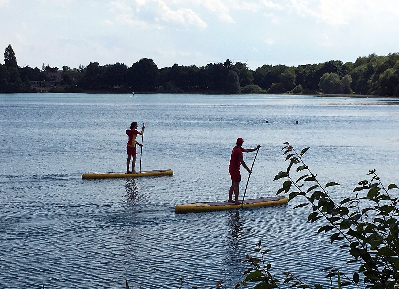 Zwei DLRG-Rettungsschwimmer patrouillieren auf Stand-Up-Paddling-Boards den Badebereich eines bayerischen Sees.