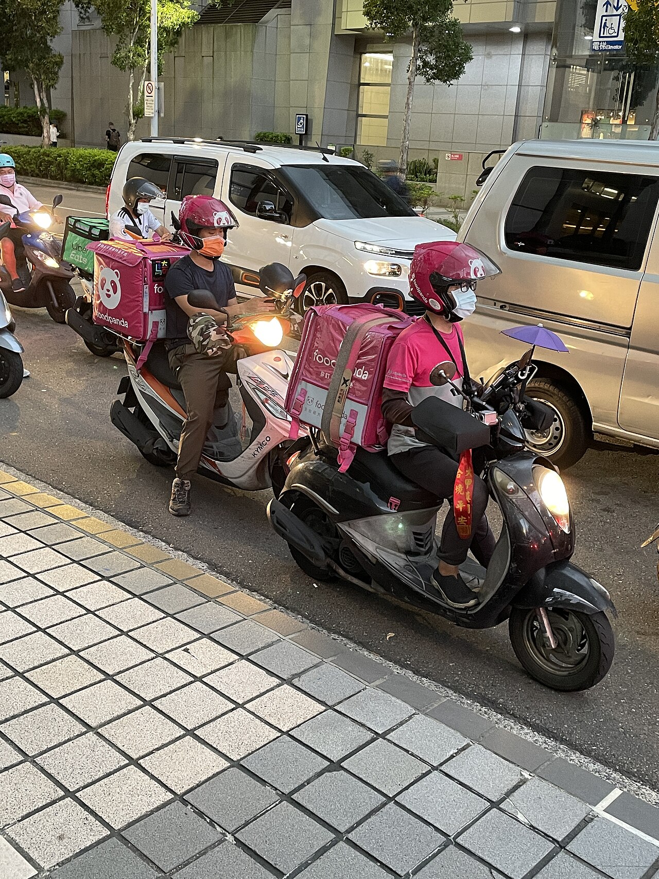 Zwei Essenskuriere von Foodpanda und Uber Eats warten an einer roten Ampel vor der U-Bahn-Station Taipei Arena.
